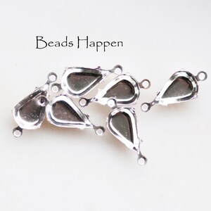 May include: Silver-toned teardrop-shaped jewelry findings with a loop at each end. The findings have a recessed area for setting a stone or other embellishment. The text "Beads Happen" is in the image.