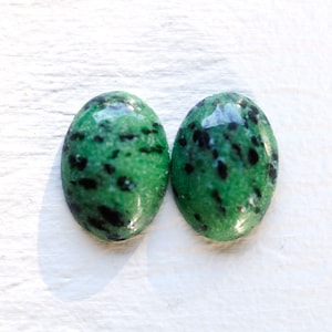 May include: Two oval-shaped green gemstones with black flecks. The stones are smooth and polished.