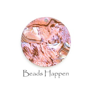 May include: A round, iridescent pendant with a hole at the top. The pendant has a swirling pattern of pink, brown, and white colors. The text "Beads Happen" is written in black script below the pendant.