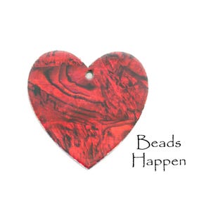 Puede incluir: Un colgante de madera en forma de corazón con un patrón veteado rojo y negro. El colgante tiene un pequeño agujero en la parte superior para ensartar. Las palabras "Beads Happen" están escritas en escritura negra debajo.