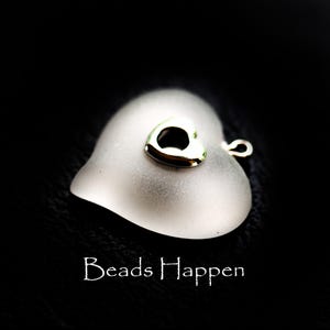 May include: A frosted glass heart pendant with a small, gold-colored heart charm on top. The pendant has a silver-colored loop for a necklace. The words "Beads Happen" are written in white script at the bottom of the image.