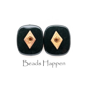 Op de afbeelding: Twee zwarte vierkante knoppen met een gouden ruitvormig ontwerp in het midden. De knoppen hebben een gat in het midden van de ruit. De tekst "Beads Happen" is onder de knoppen gedrukt.