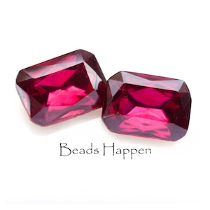 May include: Two deep red, faceted, rectangular glass beads. The beads are on a white surface.