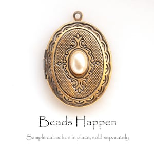 May include: A gold oval locket with a decorative design and a white pearl cabochon in the centre. The locket has a hinge on the side and a loop at the top for attaching a chain. The text "Beads Happen" and "Sample cabochon in place, sold separately" is printed below the locket.