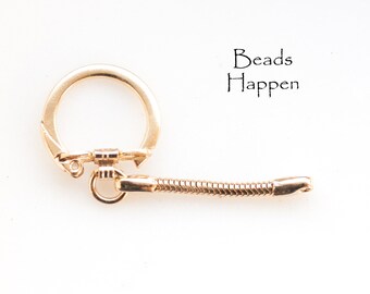 Puede incluir: Un llavero dorado con un anillo redondo y una cadena flexible. La cadena está unida al anillo con un pequeño cierre dorado. El texto "Beads Happen" está impreso sobre un fondo blanco.