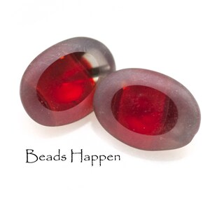 May include: Two oval-shaped glass beads with a red centre and a translucent grey outer layer. The beads are slightly different sizes and are sitting on a white surface.