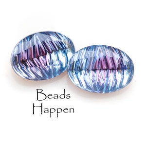 Può includere: Due perline ovali in vetro iridescente con uno schema di colori blu e viola. Le perline hanno un motivo striato e una superficie riflettente. Il testo "Beads Happen" è stampato sotto le perline.