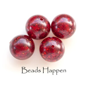 May include: Four red round beads with a glittery finish. The beads have a hole in the center for stringing. The beads are on a white background. The text "Beads Happen" is written below the beads.