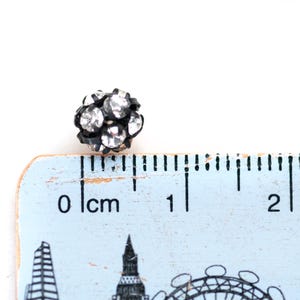 May include: A small, black and silver rhinestone ball measuring 1 cm in diameter. The ball is sitting on a ruler with markings in centimetres.