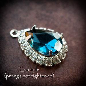 May include: A teardrop-shaped blue rhinestone pendant with a silver setting and a halo of smaller rhinestones. The prongs are not tightened.