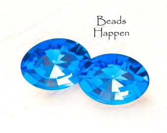 May include: Two oval-shaped, bright blue, faceted glass beads. The beads are on a white surface. The text "Beads Happen" is in the upper right corner of the image.