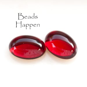 Op de afbeelding: Twee rode glazen kralen, waarvan er één iets groter is dan de andere, op een witte achtergrond. De tekst "Beads Happen" staat boven de kralen.