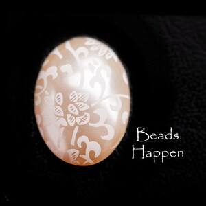 May include: An oval-shaped, peach-colored bead with a white floral design. The bead has a glossy finish and is set against a black background. The text "Beads Happen" is in white.