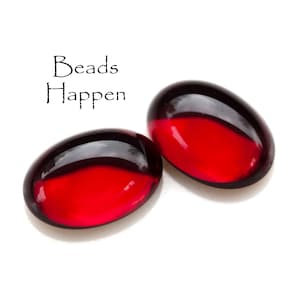 Op de afbeelding: Twee rode glazen kralen met een glanzende afwerking. De kralen zijn ovaalvormig en hebben een donkerrode kleur aan de bovenkant en een lichtere rode kleur aan de onderkant. De tekst "Beads Happen" is in zwart boven de kralen geschreven.