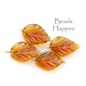May include: Four amber glass leaf-shaped beads with gold-toned metal loops. The beads have a textured surface and are arranged on a white background. The text "Beads Happen" is visible in the upper right corner of the image.