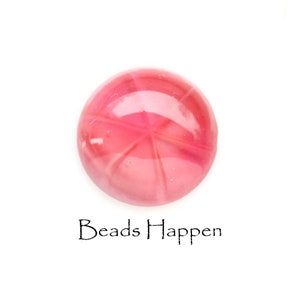 Puede incluir: Una piedra preciosa cabujón en forma de estrella rosa con una superficie lisa y pulida. La piedra preciosa está engastada en un engaste redondo transparente.