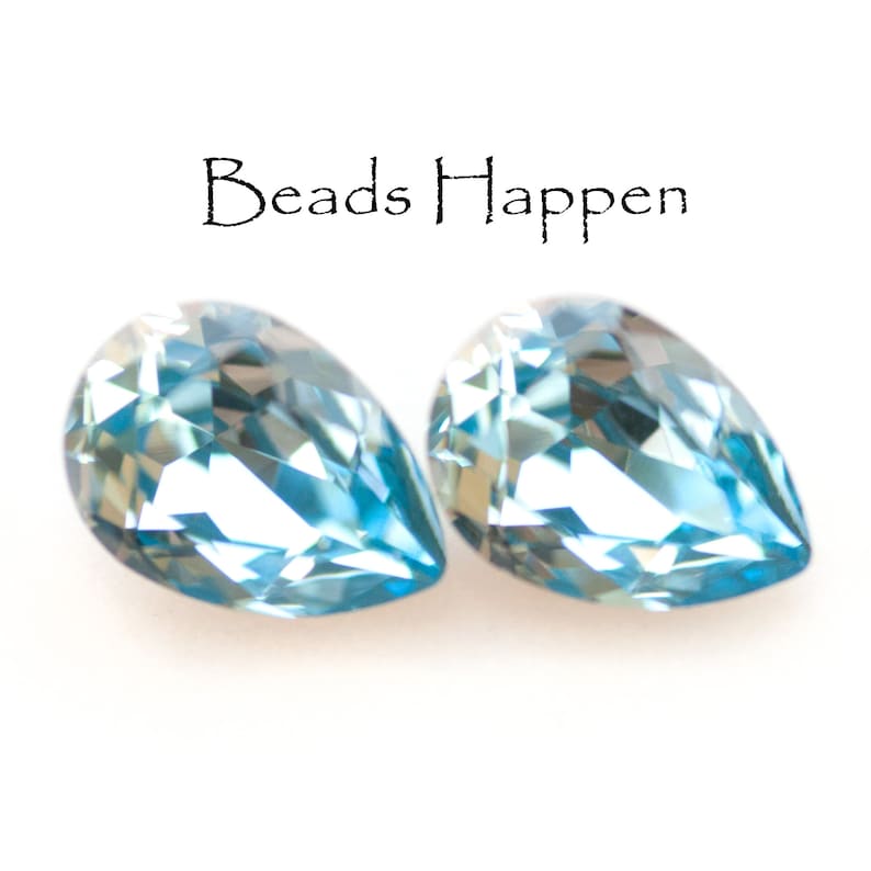 May include: Two light blue, teardrop-shaped beads with multiple facets, reflecting light. The beads are set against a white background. The text "Beads Happen" is above the beads.