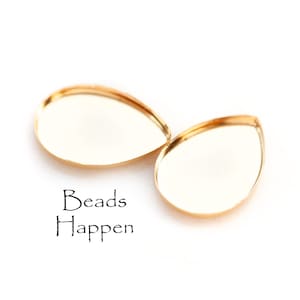 May include: Two teardrop-shaped, gold-colored bezel settings with a reflective surface. The text "Beads Happen" is printed in black script below the settings. These settings are ideal for jewelry making.