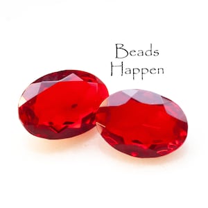Può includere: Due perline di vetro ovali rosse con un design sfaccettato. Le perline sono su uno sfondo bianco. Il testo "Beads Happen" è scritto in nero sopra le perline.