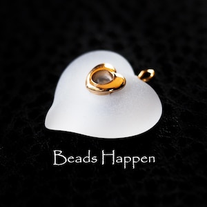 May include: A frosted glass heart pendant with a small gold heart charm on top. The pendant has a gold loop for hanging. The text "Beads Happen" is written in white script at the bottom of the image.