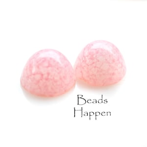 May include: Two pink, domed beads with a marbled appearance. The beads are set against a white background. The text "Beads Happen" is written in black script below the beads.