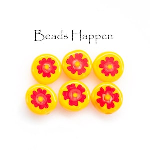May include: Six yellow glass beads with red flower designs. The beads have a flat, round shape and are arranged in two rows of three. The text "Beads Happen" is above the beads.