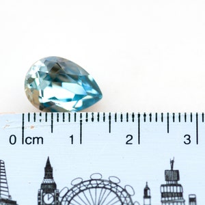 May include: A faceted, pear-shaped gemstone in shades of blue and teal. The gem is positioned next to a ruler, with measurements in centimeters. The gem is approximately 1 inch in length.