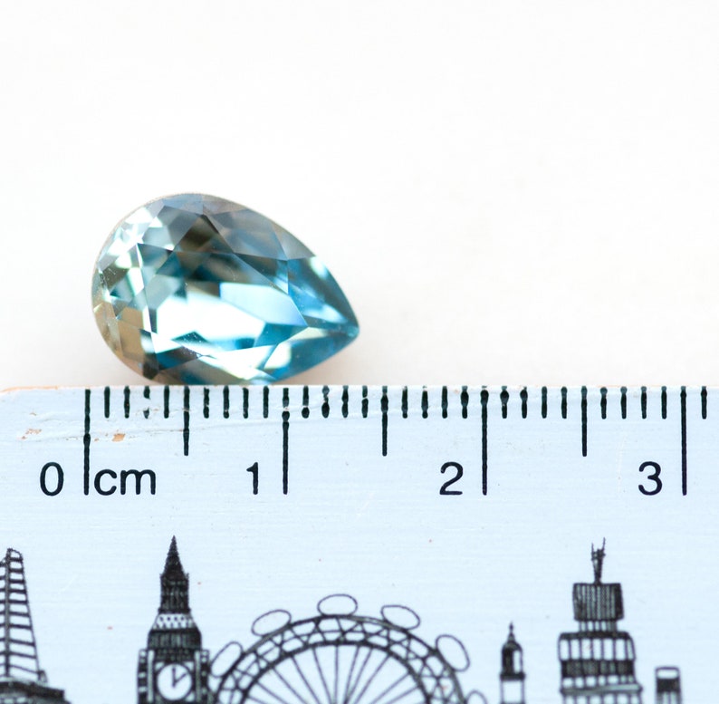 May include: A faceted, pear-shaped gemstone in shades of blue and teal. The gem is positioned next to a ruler, with measurements in centimeters. The gem is approximately 1 inch in length.