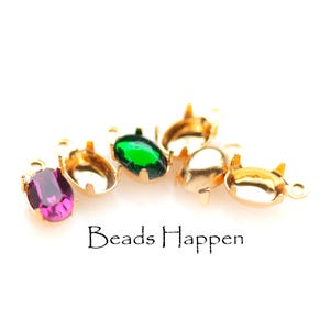 May include: Close-up of gold-coloured metal jewellery findings with oval-shaped, colourful gemstones. The gemstones are purple and green. The text "Beads Happen" is printed in black.