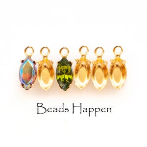 May include: A collection of gold-colored jewelry findings, including a marquise-shaped iridescent crystal and a green crystal. The findings have a loop at the top for attaching to other jewelry components. The text "Beads Happen" is printed below the findings.