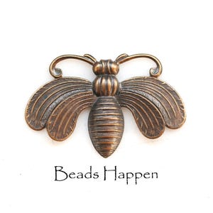 May include: A bronze-toned metal bee embellishment featuring detailed wing textures and a segmented body. The insect-shaped piece has curved antennae and is positioned above the black text that reads Beads Happen on a plain white background.