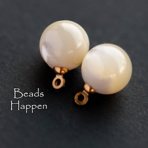 May include: Two white pearl beads with gold jump rings. The beads are round and smooth. The jump rings are attached to the top of the beads.