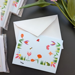 May include: Light blue greeting cards with a floral and insect design. The cards feature illustrations of flowers, a bee, a ladybug, and a snail. White envelopes are included. A pink flower and green leaves are in the background.