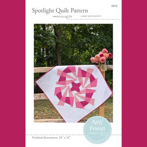 Puede incluir: Un patrón de acolchado en patchwork llamado "Spotlight" con un diseño geométrico rosa y blanco. La colcha mide 34 pulgadas por 34 pulgadas y se muestra en una valla de madera con un ramo de flores rosas en el fondo. El patrón es de Amy Friend.