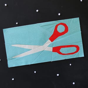 May include: A fabric art piece depicting red scissors on a light blue background with white polka dots. The scissors' blades are white, and the handles are red. The artwork is set against a dark background with small white accents.