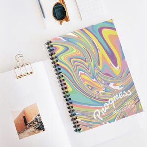 Puede incluir: Un cuaderno con espiral con un diseño colorido y arremolinado en tonos de azul, amarillo, rosa y morado. La portada presenta las palabras "Progress Over Perfection" en escritura blanca. El cuaderno está abierto, revelando páginas en blanco.