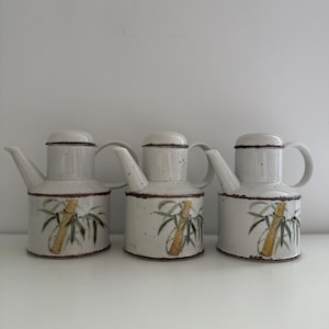 Puede incluir: Tres jarras de cerámica con tapas y asas. Las jarras son de color blanquecino con motas marrones y un diseño de bambú en verde y amarillo. Las jarras tienen un pico para verter.