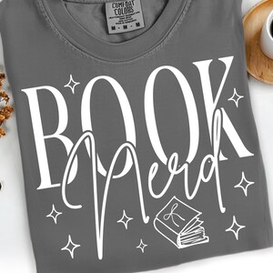 May include: A gray t-shirt with the words "BOOK Nerd" in white script. The design includes star accents and a small book illustration. The shirt is made by Comfort Colors.
