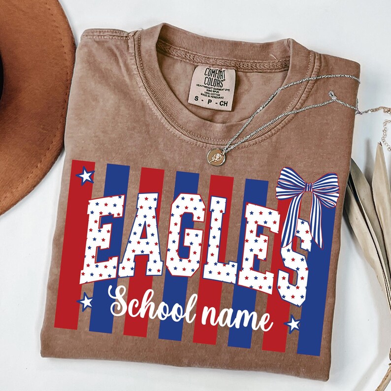 May include: A brown t-shirt with the word "EAGLES" in white, filled with red and blue stars. Red and blue stripes are behind the word. A bow graphic and the words "School name" are also on the shirt. A silver necklace is draped across the shirt.