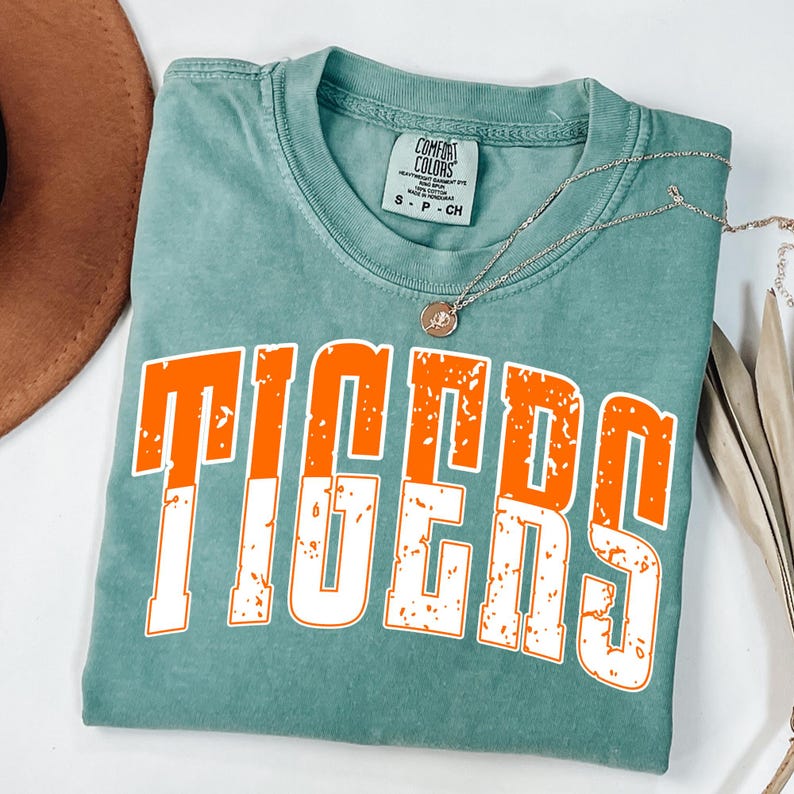 May include: A teal Comfort Colors t-shirt with the word "TIGERS" in large orange letters with a white outline. A silver necklace with a small pendant is draped across the shirt. A brown hat is visible in the upper left corner.