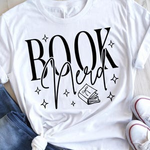 May include: White t-shirt with the words "BOOK Nerd" in black script and a small book illustration. The design includes star accents. The shirt is paired with blue jeans and white sneakers.