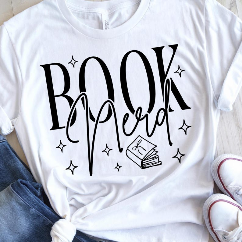 May include: White t-shirt with the words "BOOK Nerd" in black script and a small book illustration. The design includes star accents. The shirt is paired with blue jeans and white sneakers.