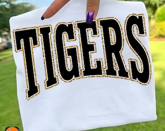 May include: White t-shirt with the word "TIGERS" in large black letters outlined with gold glitter. The shirt is held up by a hand with purple nail polish. The bottom of the image has the text "TIGERS PNG" in white on an orange background.