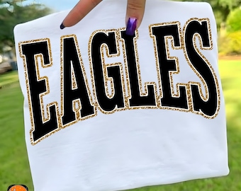May include: White t-shirt with the word "EAGLES" in large black block letters outlined with gold glitter. The shirt is held up by a hand with purple nail polish. The bottom of the shirt has the text "EAGLES PNG".