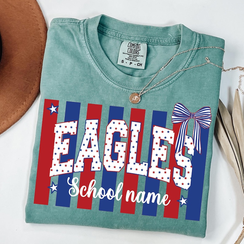 May include: A seafoam green t-shirt with a patriotic design. The word "EAGLES" is printed in white with red and blue star accents. Below, the words "School name" are in a script font. A bow with red, white, and blue stripes is on the right.
