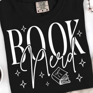 May include: Black t-shirt with the words "BOOK Nerd" in white script and block letters. The design includes a small book graphic and star shapes. The shirt is made by Comfort Colors.