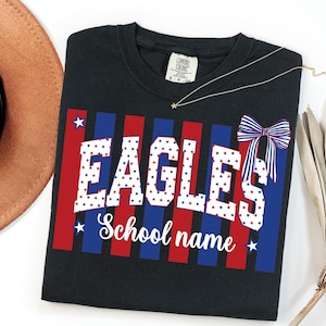 May include: Black t-shirt with the word "EAGLES" in white, red, and blue star-filled letters. The shirt has red and blue vertical stripes and a bow graphic. The words "School name" are below the word "EAGLES".