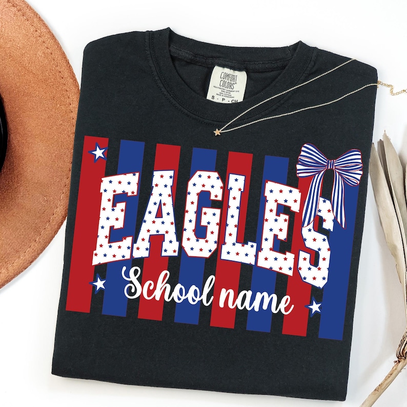May include: Black t-shirt with the word "EAGLES" in white, red, and blue star-filled letters. The shirt has red and blue vertical stripes and a bow graphic. The words "School name" are below the word "EAGLES".