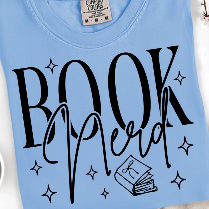 May include: Light blue t-shirt with the words "BOOK Nerd" in black script and a small book illustration. The shirt also features small star shapes around the text. The shirt is made of a soft material.