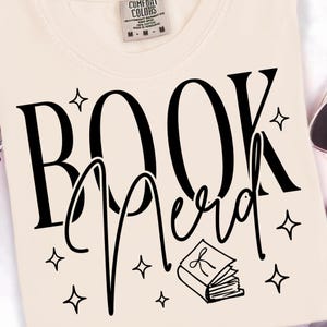 May include: Cream-colored sweatshirt with the words "BOOK Nerd" in large black letters. The word "Nerd" is in a script font. The design includes star shapes and a small book illustration.